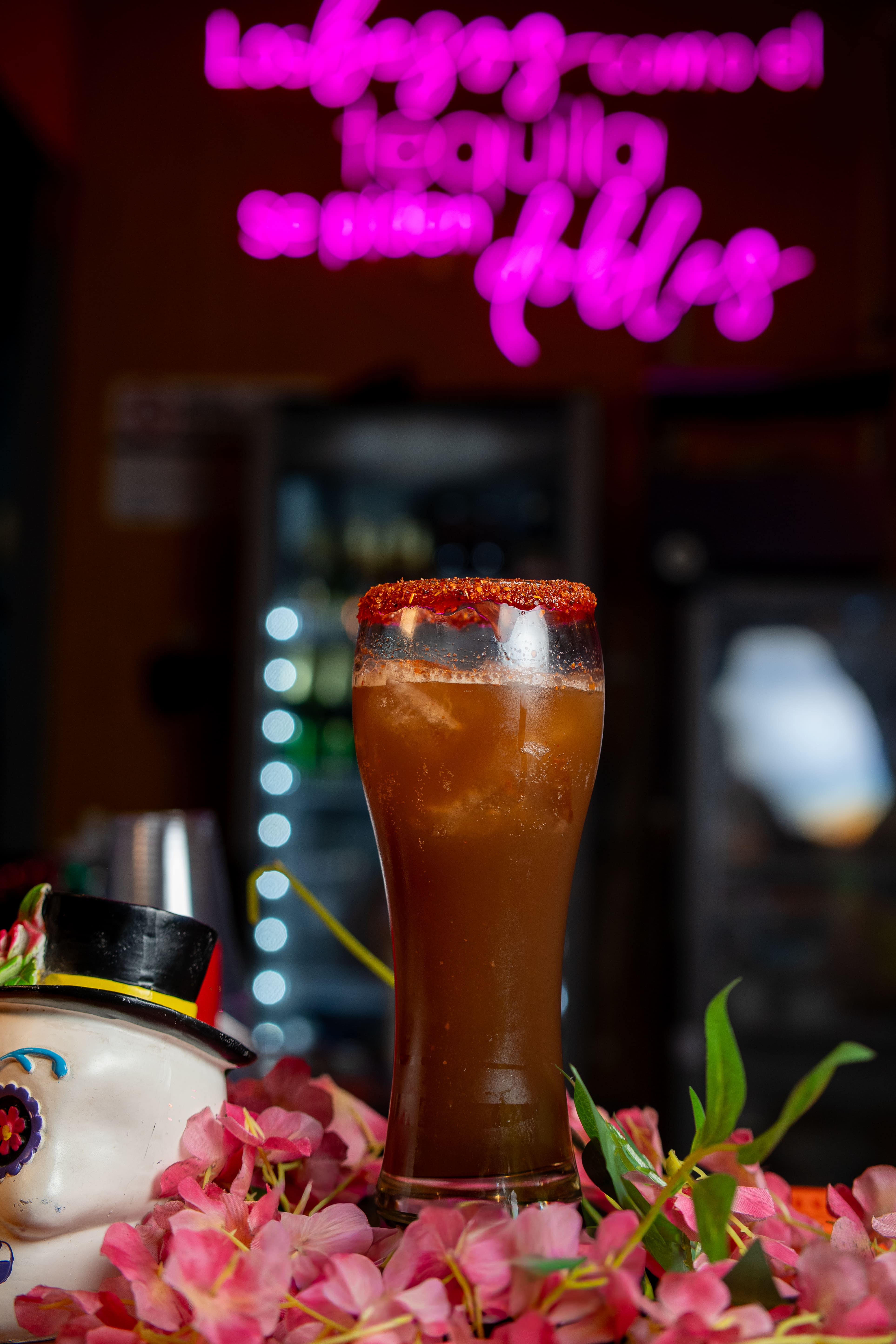 Fotografía de barra que destaca un vaso largo con una michelada o bebida oscura escarchada con chile en polvo