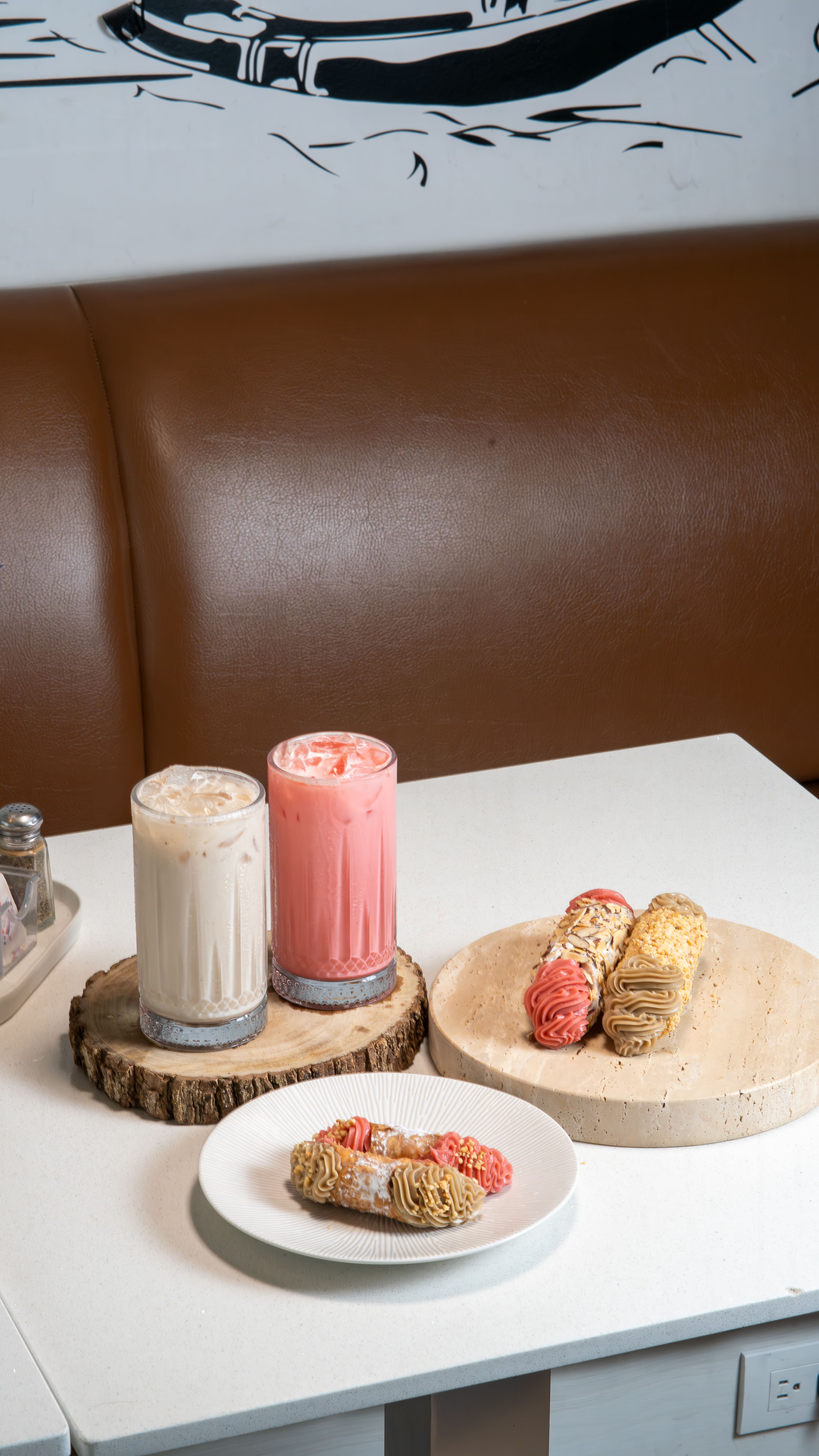 Una toma limpia con luz difusa que presenta un bodegón de postres sobre una mesa blanca: dos vasos con bebidas frías , un plato con cannolis rellenos de crema de fresa y chocolate, y otra base con más piezas del mismo postre italiano